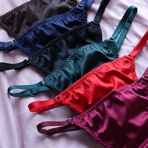 Può includere: Una collezione di sei bralette in raso disposte in fila diagonale, con colori che includono marrone scuro, blu navy, nero, verde smeraldo, rosso brillante e magenta intenso. Ogni pezzo presenta bordi in tessuto arricciato e spalline regolabili.