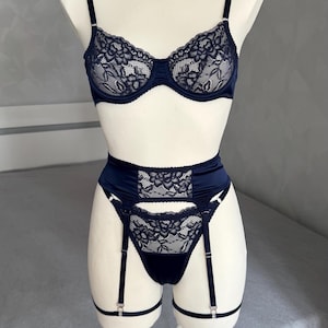 May include: A dark navy blue lingerie set consisting of an underwire bra and a high-waisted garter belt with matching thong. The set features intricate black floral lace overlays on a smooth satin fabric, with adjustable straps and metal hardware.