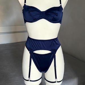 May include: Navy blue lingerie set. The set comprises a bra with underwire support, a high-waisted suspender belt, and matching briefs. The lingerie is made of a smooth, satin-like fabric. The suspender belt has adjustable straps and metal hardware.