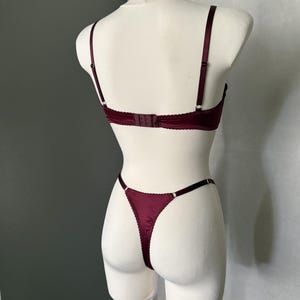 May include: A deep burgundy lingerie set consisting of a bra and a thong. The bra features thin adjustable shoulder straps and a back hook-and-eye closure. The matching thong has thin side straps and a scalloped edge trim throughout the set.