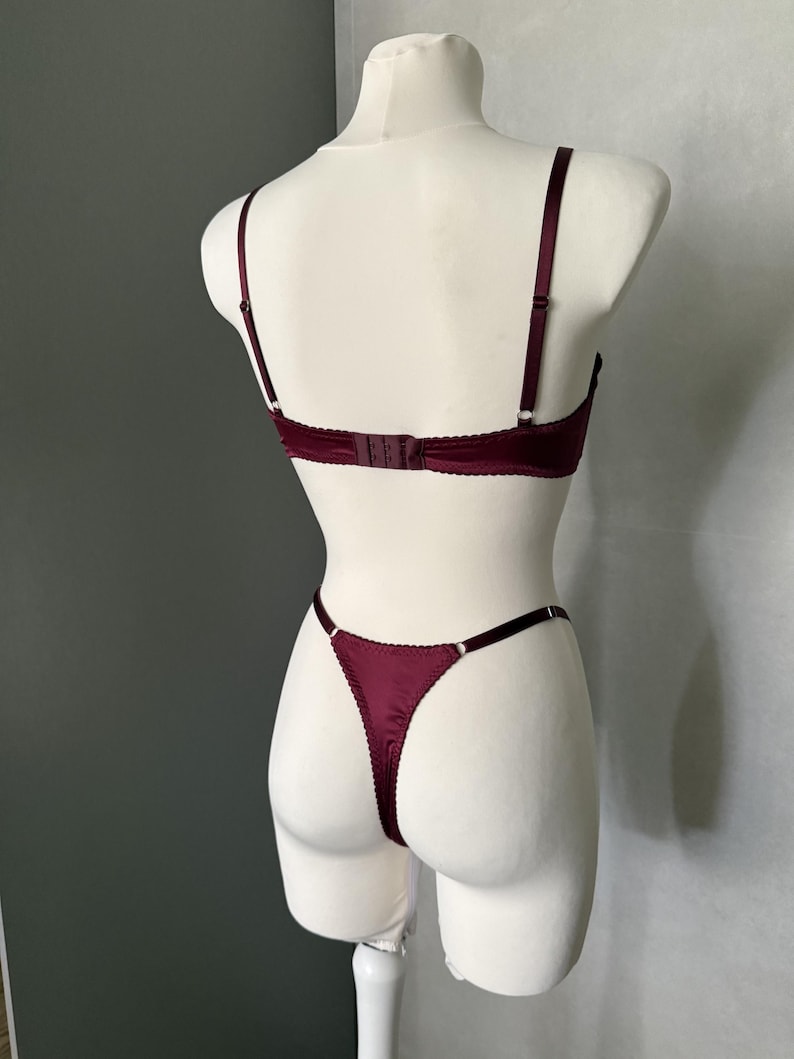 May include: A deep burgundy lingerie set consisting of a bra and a thong. The bra features thin adjustable shoulder straps and a back hook-and-eye closure. The matching thong has thin side straps and a scalloped edge trim throughout the set.