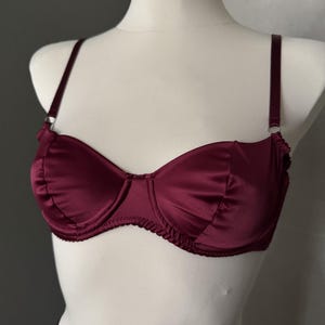 May include: A deep burgundy satin bra featuring underwire cups with gathered fabric detailing along the edges, thin adjustable shoulder straps with silver-toned metal rings, and a matching burgundy fabric band with a scalloped trim finish.