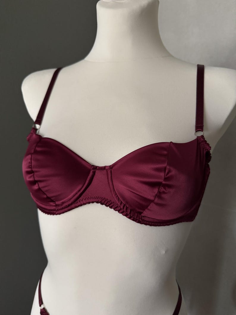 May include: A deep burgundy satin bra featuring underwire cups with gathered fabric detailing along the edges, thin adjustable shoulder straps with silver-toned metal rings, and a matching burgundy fabric band with a scalloped trim finish.