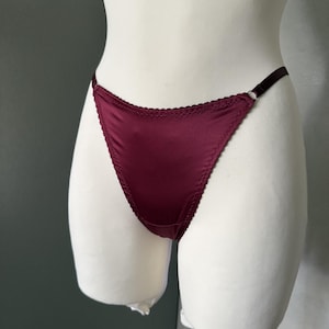 May include: A deep burgundy satin thong featuring a high-cut leg design with delicate scalloped edges along the waistband and leg openings. The garment includes thin, adjustable elastic side straps connected by small metal rings for a secure fit.
