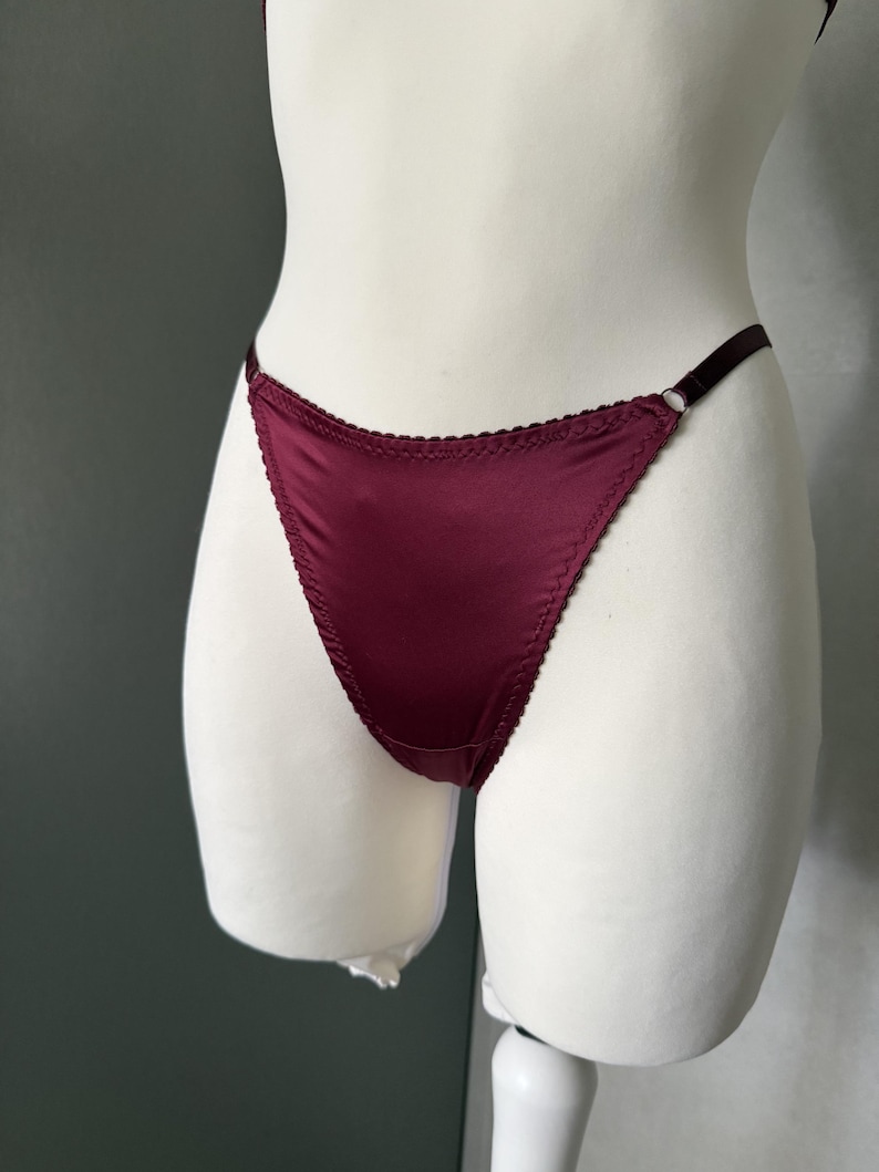 May include: A deep burgundy satin thong featuring a high-cut leg design with delicate scalloped edges along the waistband and leg openings. The garment includes thin, adjustable elastic side straps connected by small metal rings for a secure fit.
