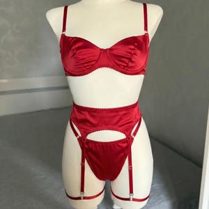 May include: A vibrant red lingerie set featuring a satin underwire bra with adjustable shoulder straps, paired with a high-waisted suspender belt and matching knickers. The set includes attached suspender straps with metal clips for hosiery.