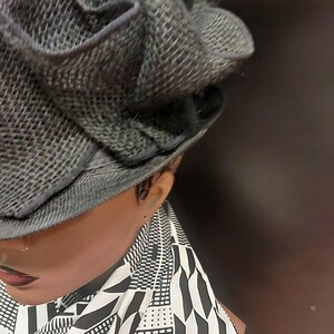 May include: A grey, woven hat with a large, ruffled brim. The hat is worn by a person with a black and white patterned shirt.