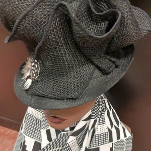 May include: A black, wide-brimmed hat with a large, ruffled crown made of woven fabric. The hat is decorated with a small, black and white feathered ornament.