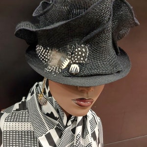 May include: A black, wide-brimmed hat with a ruffled crown and a black and white feather accent. The hat is worn by a mannequin with a black and white patterned scarf.