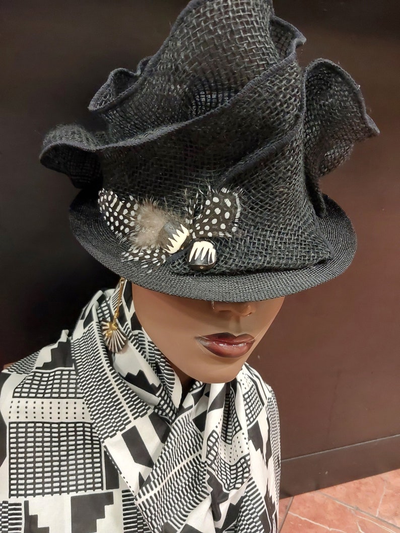 May include: A black, wide-brimmed hat with a ruffled crown and a black and white feather accent. The hat is worn by a mannequin with a black and white patterned scarf.