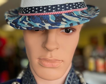 May include: A white fedora hat with a blue and white camouflage patterned brim. The hat features a band with a pattern of white stars and red stripes. The hat is displayed on a mannequin.