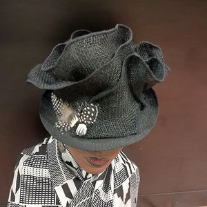 May include: A black, ruffled, straw hat with a small feather accent. The hat is worn on a mannequin with a black and white patterned shirt.