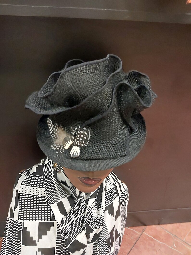 May include: A black, ruffled, straw hat with a small feather accent. The hat is worn on a mannequin with a black and white patterned shirt.