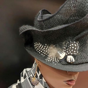 May include: A black and white patterned hat with a feather and a decorative pin. The hat is worn by a mannequin with gold earrings. The mannequin is wearing a black and white patterned scarf.