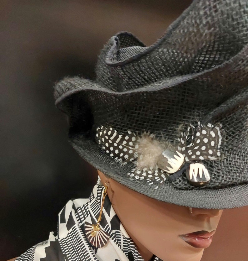 May include: A black and white patterned hat with a feather and a decorative pin. The hat is worn by a mannequin with gold earrings. The mannequin is wearing a black and white patterned scarf.