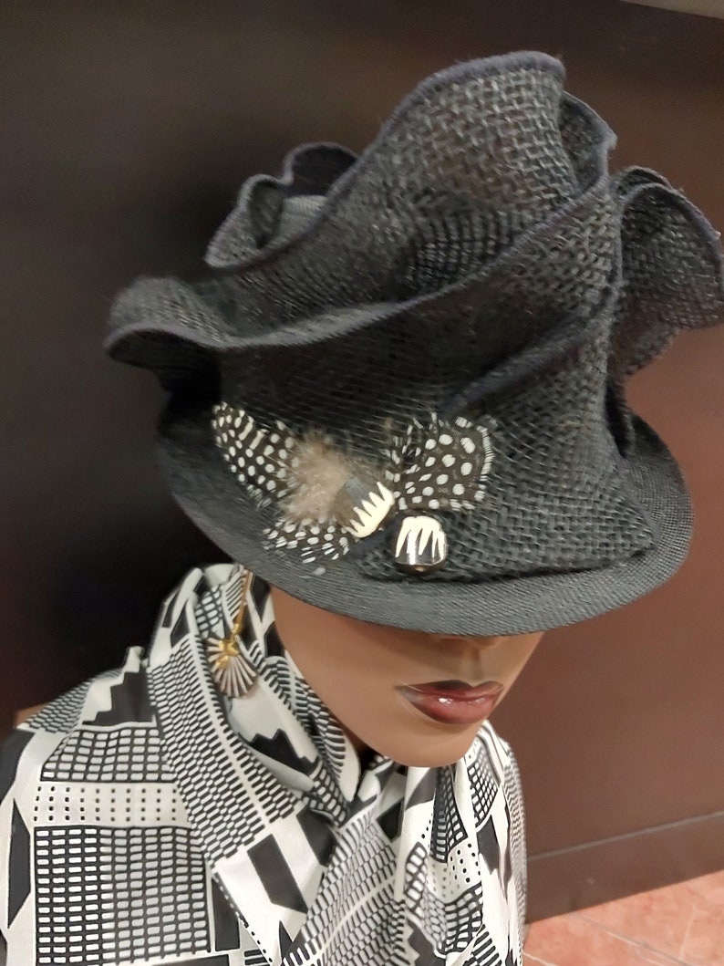 May include: A black, ruffled hat with a white and black feather accent. The hat is worn on a mannequin with a black and white patterned scarf.