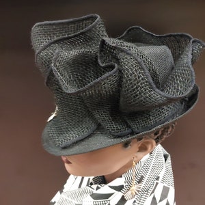 May include: A black mesh hat with a wide brim and ruffled detailing. The hat is worn by a person with a black and white patterned scarf and gold earrings.