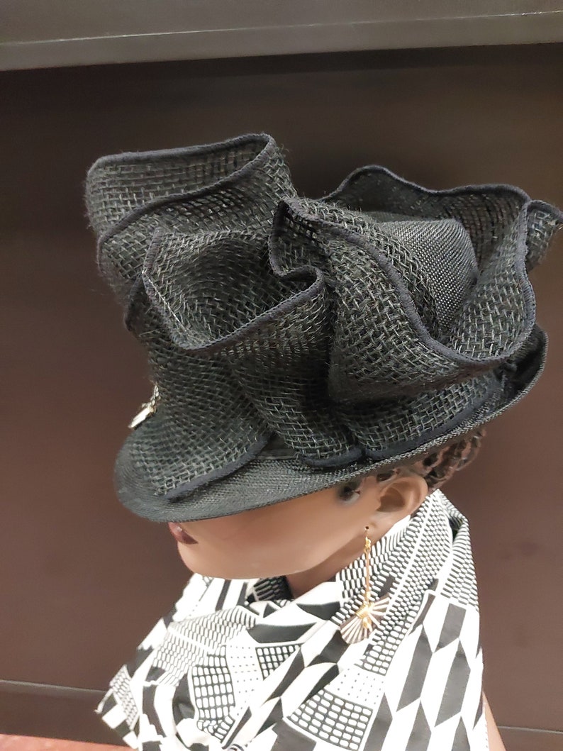May include: A black mesh hat with a wide brim and ruffled detailing. The hat is worn by a person with a black and white patterned scarf and gold earrings.