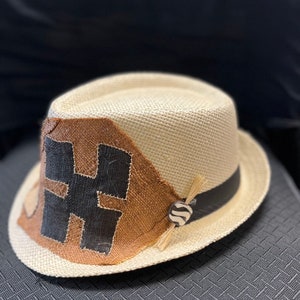 May include: A fedora-style hat with a natural straw-like weave. The hat features a brown patch with a black "H" design, a black band, and a decorative button with a zebra pattern. A small bundle of straw-like material is attached to the button.