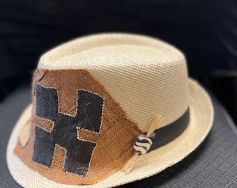 May include: A fedora-style hat with a natural straw-like weave. The hat features a brown patch with a black "H" design, a black band, and a decorative button with a zebra pattern. A small bundle of straw-like material is attached to the button.
