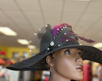 May include: A black wide-brimmed hat with decorative feathers, netting, and embellishments. The hat is adorned with white beads and shell accents. The mannequin is wearing a colorful patterned top.