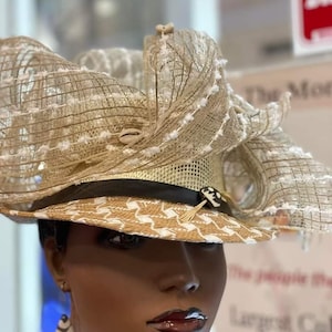 May include: A beige and white woven hat with a wide brim and decorative accents. The hat features a black band, a button with a tribal design, and a layered, textured crown. The brim has a geometric pattern. The hat is displayed on a mannequin.