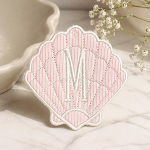 Puede incluir: Un parche en forma de concha rosa y blanca con la letra "M" bordada en blanco. El parche tiene un diseño acolchado con rayas verticales y un borde festoneado. El fondo es una superficie de mármol.