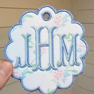 Op de afbeelding: Een geschulpt, rond stoffen label met een witte achtergrond en een bloemenpatroon in lichtblauw, roze en groen. Het midden is geborduurd met de lichtblauwe monogramletters JHM in een schreeflettertype en de rand is afgewerkt met blauw stiksel.