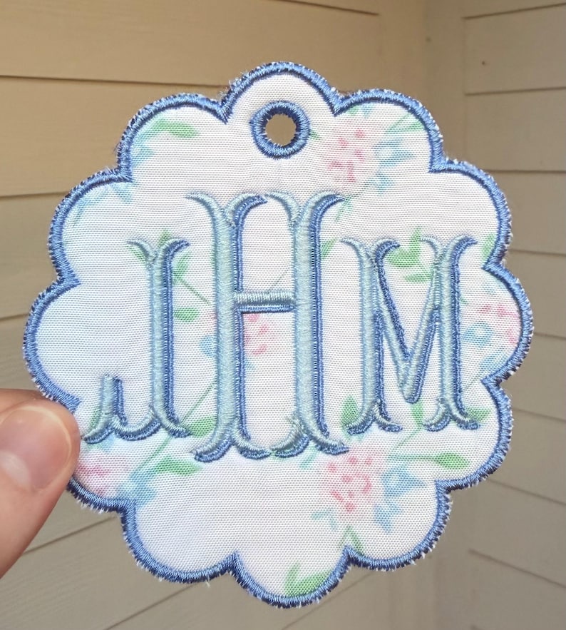Op de afbeelding: Een geschulpt, rond stoffen label met een witte achtergrond en een bloemenpatroon in lichtblauw, roze en groen. Het midden is geborduurd met de lichtblauwe monogramletters JHM in een schreeflettertype en de rand is afgewerkt met blauw stiksel.