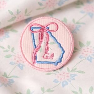 May include: A circular patch with the shape of Georgia in blue, a pink bow, and the letters "GA". The patch has a pink and white striped background and a pink border. The background is a floral pattern.