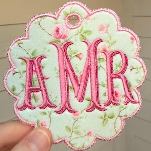 Op de afbeelding: Een geschulpt stoffen label met een lichtgroene achtergrond en een roze rozenpatroon. De letters AMR zijn in het midden geborduurd met felroze draad. De randen en een klein rond gaatje aan de bovenkant zijn afgewerkt met roze stiksels.