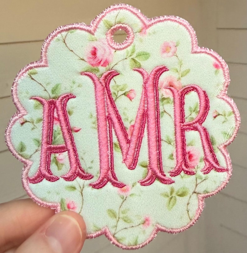 Op de afbeelding: Een geschulpt stoffen label met een lichtgroene achtergrond en een roze rozenpatroon. De letters AMR zijn in het midden geborduurd met felroze draad. De randen en een klein rond gaatje aan de bovenkant zijn afgewerkt met roze stiksels.