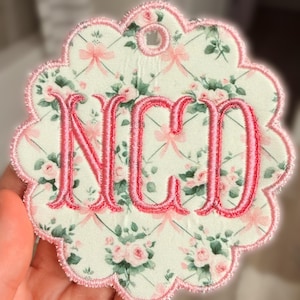 Op de afbeelding: Een cirkelvormig stoffen ornament met een geschulpte rand, voorzien van een lichtgroene achtergrond met een patroon van roze rozen, groene bladeren en dunne roze linten. Het midden toont de geborduurde roze monogramletters NCD met een rand.