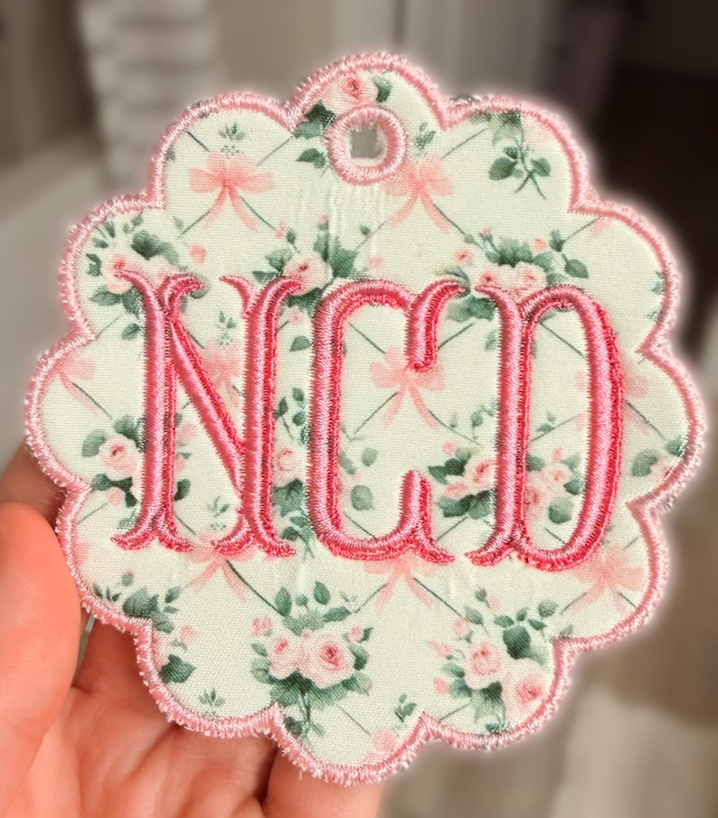 Op de afbeelding: Een cirkelvormig stoffen ornament met een geschulpte rand, voorzien van een lichtgroene achtergrond met een patroon van roze rozen, groene bladeren en dunne roze linten. Het midden toont de geborduurde roze monogramletters NCD met een rand.