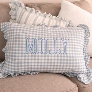Pu&ograve; includere: Un cuscino rettangolare con motivo a quadretti vichy azzurro e bianco e bordo arricciato. Il nome MOLLY &egrave; ricamato al centro con un carattere serif blu. Il cuscino &egrave; posizionato davanti ad altri cuscini.