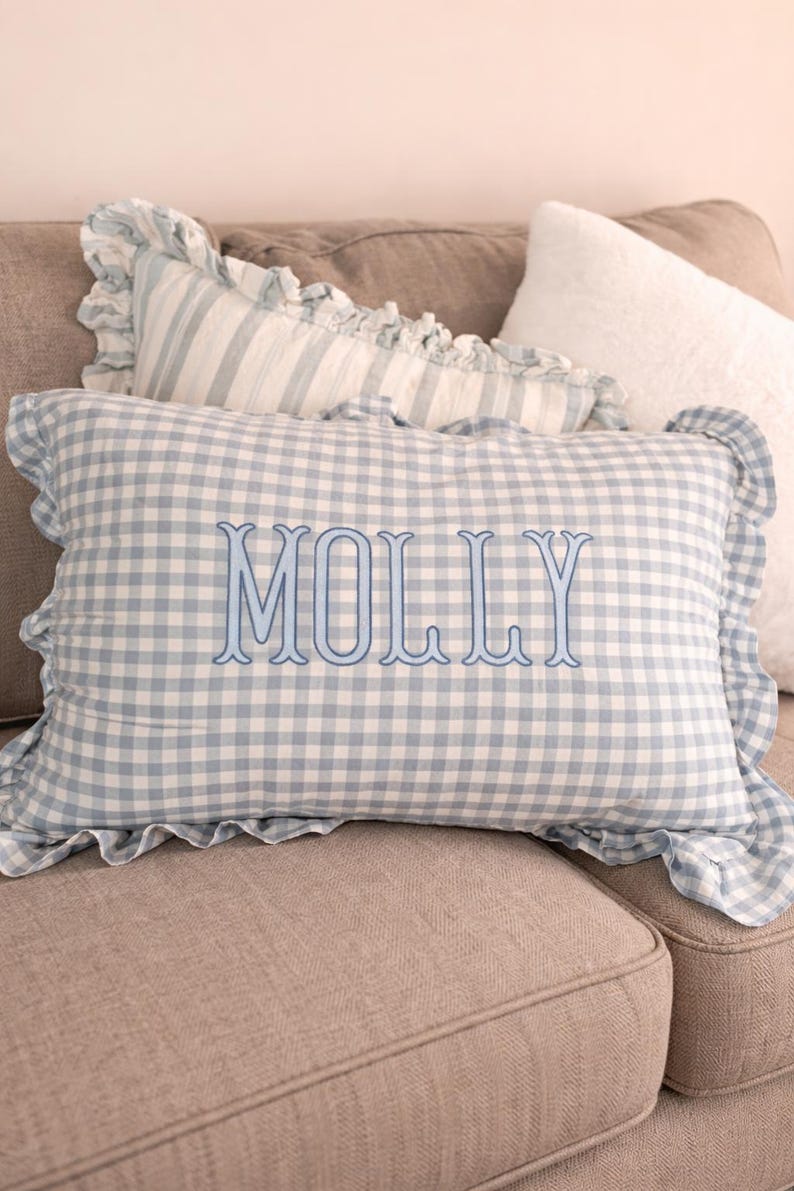 Pu&ograve; includere: Un cuscino rettangolare con motivo a quadretti vichy azzurro e bianco e bordo arricciato. Il nome MOLLY &egrave; ricamato al centro con un carattere serif blu. Il cuscino &egrave; posizionato davanti ad altri cuscini.
