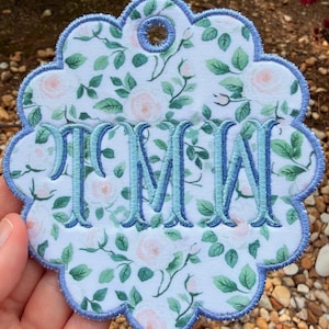 Op de afbeelding: Een cirkelvormig stoffen ornament met een geschulpte rand, voorzien van een witte achtergrond met een patroon van lichtroze rozen en groene bladeren. Het midden is geborduurd met de blauwe monogramletters TMW en de randen zijn blauw afgewerkt.