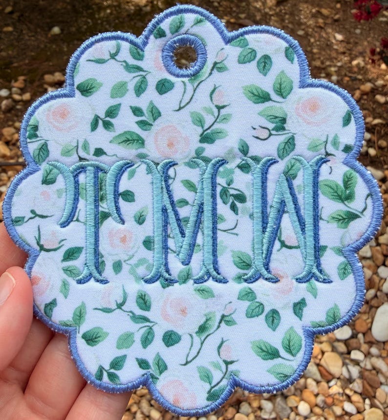 Op de afbeelding: Een cirkelvormig stoffen ornament met een geschulpte rand, voorzien van een witte achtergrond met een patroon van lichtroze rozen en groene bladeren. Het midden is geborduurd met de blauwe monogramletters TMW en de randen zijn blauw afgewerkt.