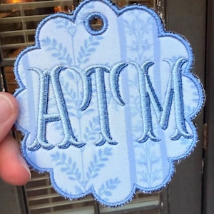 Op de afbeelding: Een lichtblauw geschulpt stoffen label met een donkerblauw geborduurd monogram van de letters ATM. De stof heeft een subtiel verticaal streeppatroon met delicate bladmotieven. Een cirkelvormig gat is in het midden bovenaan om op te hangen.