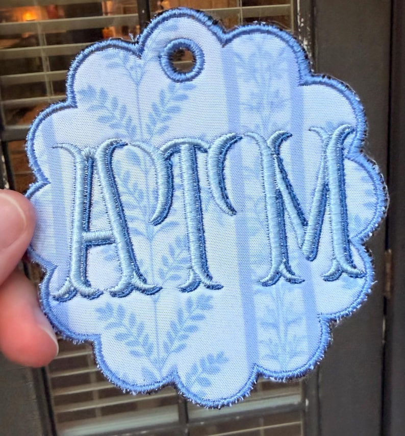 Op de afbeelding: Een lichtblauw geschulpt stoffen label met een donkerblauw geborduurd monogram van de letters ATM. De stof heeft een subtiel verticaal streeppatroon met delicate bladmotieven. Een cirkelvormig gat is in het midden bovenaan om op te hangen.