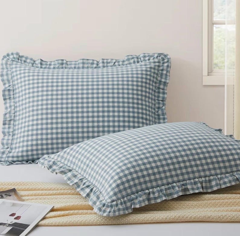 Pu&ograve; includere: Due cuscini rettangolari con un motivo a quadretti vichy blu e bianchi. Ogni cuscino &egrave; rifinito con un bordo in tessuto arricciato lungo tutti e quattro i lati, creando una cornice decorativa attorno al design a quadretti centrale.