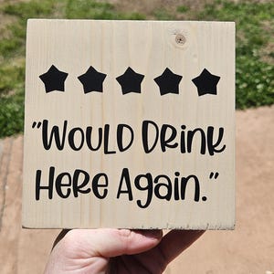 May include: A wooden sign with the phrase "Would Drink Here Again" in black lettering. Above the text are five black star shapes. The sign is held up against a blurred green background.