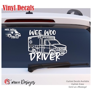 May include: A white vinyl decal applied to a car rear window featuring a line art illustration of an ambulance with the text WEE WOO DRIVER above and below the vehicle. A smaller version of the ambulance graphic is positioned to the left.