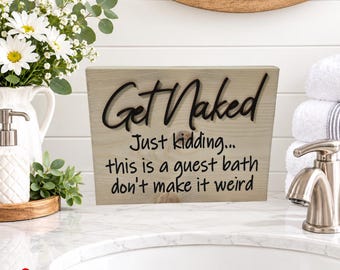 May include: A rectangular wooden sign with a light natural finish featuring black raised lettering that reads: Get Naked Just kidding... this is a guest bath don't make it weird. The sign is positioned on a white countertop near a bathroom sink.