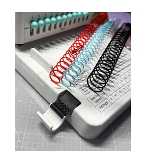 May include: A white plastic binding machine featuring a row of light blue pegs, a black fabric strap, and a printed chart for book sizes in inches. Three plastic binding coils in red, light blue, and black are positioned on the machine surface.