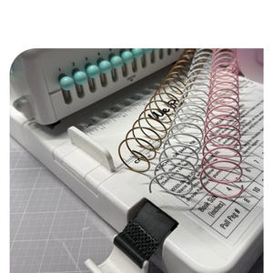 May include: A white binding machine with teal knobs and a black strap. Several metal spiral binding coils in gold, silver, and pink are arranged on the machine's surface. The machine has a ruler and text that reads "INCH OR SMALLER".