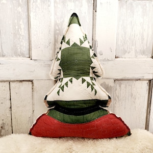 May include: A quilted Christmas tree-shaped pillow. The tree features a cream-colored background with green and red accents. The design includes green triangles and a red base, creating a festive holiday decoration.