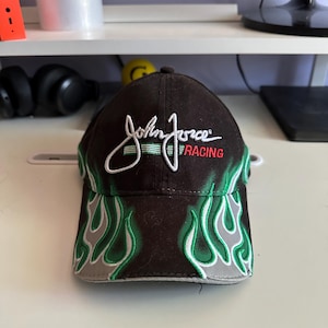 May include: A black baseball cap featuring green and grey flame patterns on the brim and front panels. The text John Force is embroidered in white script above the word RACING in red, with a horizontal green line accenting the design.