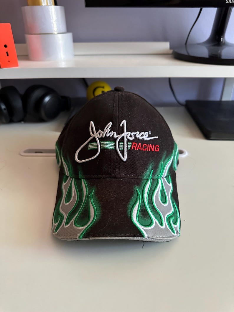 May include: A black baseball cap featuring green and grey flame patterns on the brim and front panels. The text John Force is embroidered in white script above the word RACING in red, with a horizontal green line accenting the design.