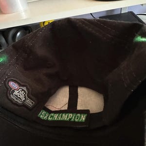 May include: A dark brown baseball cap featuring an embroidered NHRA Full Throttle Energy Drink logo on the side and a green embroidered patch on the back strap that reads 15x CHAMPION in capital letters with a white interior lining.